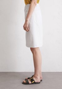 Jupe en denim blanc de forme droite, avec une poche arrière. Associée à des sandales noires avec un accent en boucle dorée.