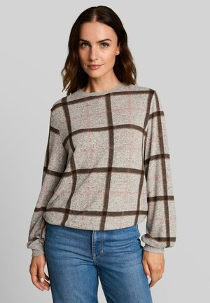Vrouw met schouderlang bruin haar, die een beige trui met lange mouwen en bruin ruitpatroon en een blauwe spijkerbroek draagt, tegen een effen grijze achtergrond.
