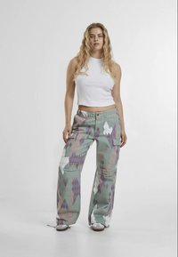Calças cargo largas e multicoloridas com padrões em losango nas cores verde, roxo e branco; apresentam bolsos e um fecho de botão à frente, combinadas com uma t-shirt sem mangas branca.