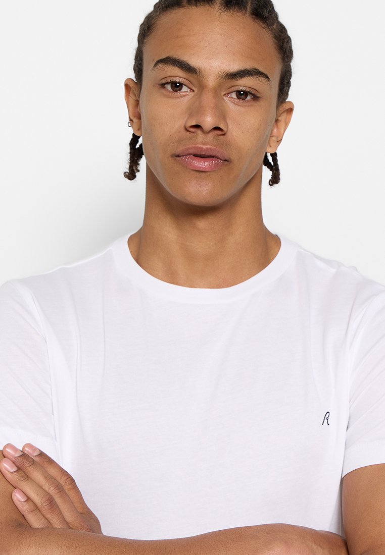 Camiseta de manga corta blanca hecha de algodón. Presenta un escote redondo y un pequeño logo oscuro en el lado izquierdo del pecho. Textura suave.