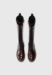 Burgundowe skórzane kozaki do kolan z sznurówkami i bocznym zamkiem; detale typu brogue i teksturowane wykończenie; płaska podeszwa dla stabilności.