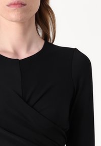 Svart langermet topp med rund halsringning og en tvistet detalj ved midjen, laget av glatt, elastisk stoff.