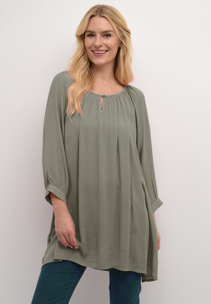 Hellgrüne, locker sitzende Bluse mit Dreiviertelärmeln, Schlüssel-Ausschnitt und strukturiertem Stoff. Kombiniert mit blauen Jeans.
