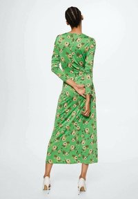 Grünes Kleid mit Blumenmuster, langen Ärmeln, tailliertem Oberteil und fließender Maxilänge. Getragen mit weißen High Heels.