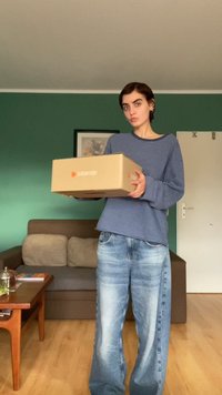 Caixa de papelão com o logótipo da Zalando em vermelho e branco, segurada por uma pessoa que veste uma camisa de manga longa azul e branca às riscas e jeans azul desbotado.