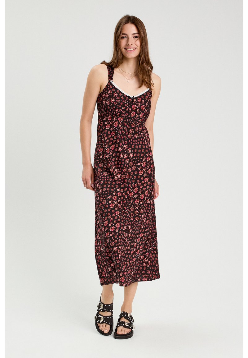 Robe midi fleurie avec un fond noir, ornée de fleurs roses, à fines bretelles et un corsage ajusté. Associée à des sandales noires à boucle.