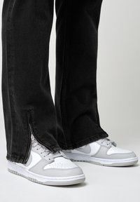 Pantalones vaqueros negros con dobladillos con cremallera, combinados con zapatillas blancas y grises con detalles perforados y suelas de goma.