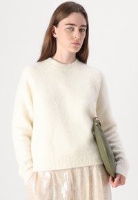 Maglione color crema, soffice, con collo a girocollo a coste, indossato sopra una gonna con paillettes. La modella tiene una borsa a tracolla verde dal design elegante.