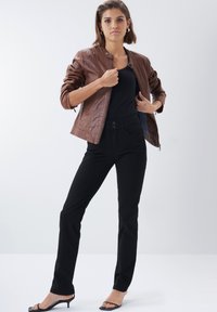 Chaqueta bomber de cuero marrón con detalles de canalé, top ajustado negro y pantalones negros, complementados con sandalias de tacón negro con tiras.