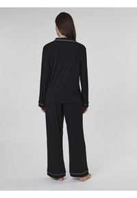 Svart pyjamasset med långa ärmar och byxor, tillverkat av mjukt tyg med vit piping längs kragen och ärmslut, sedd bakifrån.