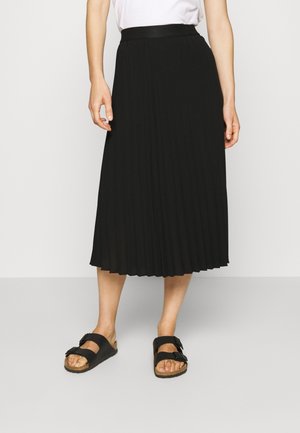 Jupe midi plissée noire avec une taille lisse, associée à des sandales noires. La jupe présente un design fluide et un tissu léger.