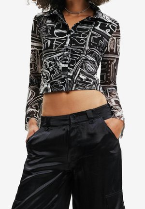 Femme portant une chemise courte boutonnée à motif abstrait noir et blanc et un pantalon large en satin noir avec les mains dans les poches.