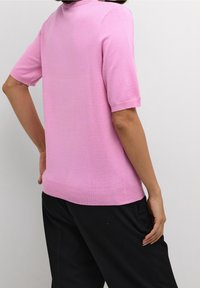 Donna che indossa un top a maniche corte rosa in maglia e pantaloni neri, in piedi con una mano sui fianchi contro uno sfondo chiaro e semplice.