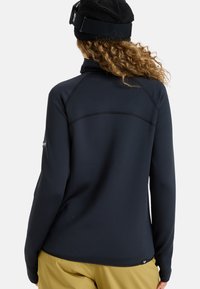 Black, textured fleece pullover with a high collar, featuring contrasting seams. Worn with light-colored pants and a dark beanie.