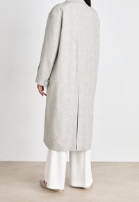 Lang grijs jack met een ruitpatroon, met een rechte snit, brede mouwen en een centrale achtersplit. Gecombineerd met witte wijde broek.