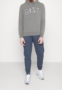 Uomo che indossa una felpa grigia GANT, pantaloni jogger blu e sneakers bianche, in piedi contro uno sfondo bianco semplice.