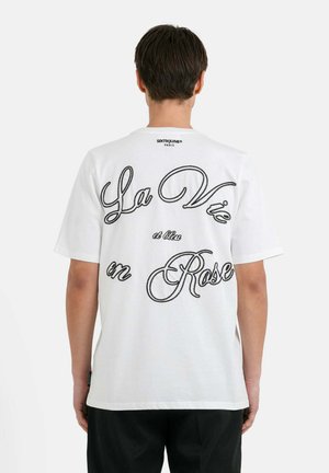 Hvid bomulds T-shirt med stor sort kursiv tekst på ryggen, der står "La Vie en Rose," med ribbet runde halsudskæring og korte ærmer.