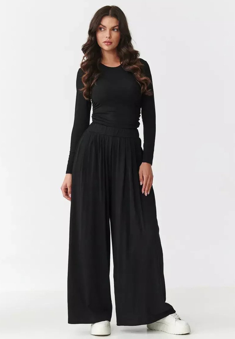 Czarna bluzka z długim rękawem w połączeniu z szerokimi czarnymi spodniami. Materiał wydaje się miękki o gładkiej fakturze. Obuwie to białe, okrągłe trampki.