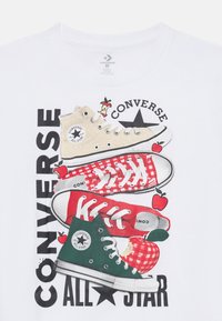 Camiseta blanca con ilustraciones de zapatillas Converse: bota alta beige, zapatilla baja de cuadros rojos y bota alta verde, con gráficos de manzanas y texto del logo.