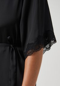 Robe en satin noir avec des manches larges, délicate dentelle, fermeture à nouer à la taille et texture lisse.