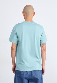 Κεφαλός άτομο που φορά ασημένια σκουλαρίκια κρίκους, ανοιχτό μπλε πουκάμισο κοντό μανίκι και μπλε τζιν, στραμμένο προς τα πίσω με τατουάζ στο δεξί του χέρι.