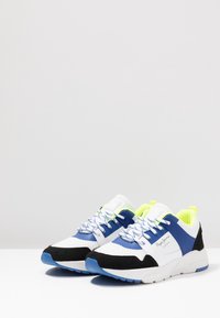 Sneaker in rete bianchi e neri con accenti blu e giallo fluorescente. Presentano pannelli a contrasto, lacci testurizzati e un colletto imbottito.
