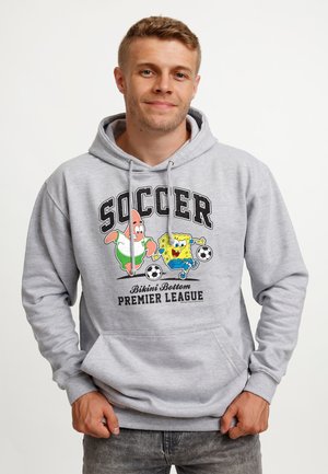 Ung mand iført en grå hættetrøje med SpongeBob og Patrick, der spiller fodbold, og teksten "Soccer Bikini Bottom Premier League."