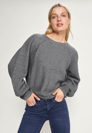 Grijze gebreide trui met een ribbelstructuur, wijde mouwen en een ronde hals. Draagt met donkerblauwe jeans, die een relaxte pasvorm laten zien.