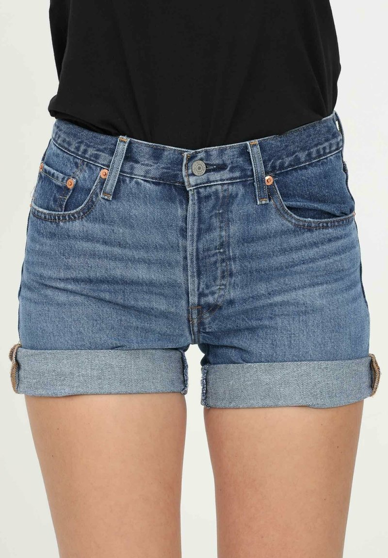 Femme portant un short en jean bleu roulé et un haut noir, montrée de la taille jusqu'à mi-cuisse.