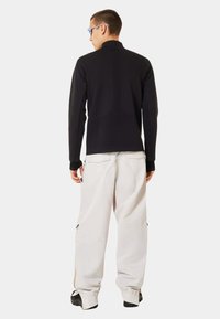 Black long-sleeve top with high collar and textured design; paired with loose-fitting, light gray pants featuring side zippers and pockets.