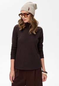 Frau mit einer beigen Strickmütze, übergroßen Schildkrötenglasen, einem braunen strukturierten Langarmoberteil, braunen Hosen und Armbändern am linken Handgelenk.