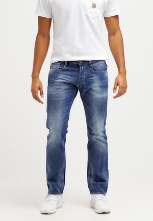Jeans de denim azul con corte recto, ligero desgastado, cinco bolsillos y detalles de costura. Llevados con una camiseta blanca y zapatillas blancas.