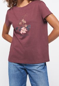 Persona que lleva una camiseta burdeos con un gráfico floral y el texto "MUSTANG est. 1932", combinada con unos jeans de mezclilla azules.