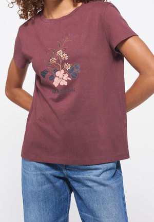 Personne portant un t-shirt bordeaux avec un motif floral et le texte "MUSTANG est. 1932", assorti à un jean en denim bleu.