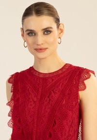 Rotes Spitzenoberteil mit aufwendigen Blumenmustern, welligen Kanten und einem hohen Kragen. Ärmelloses Design mit einer strukturierten, geschichteten Optik.