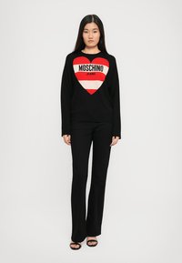 Svart genser med hjerte-design som har røde og hvite striper, med teksten "MOSCHINO JEANS". Brukt med vide svarte bukser og sandaler.