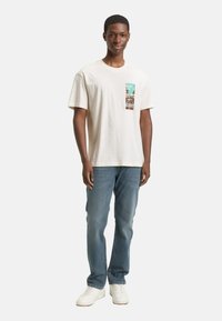 Kurzes cremefarbenes T-Shirt mit einem grafischen Brusttaschen-Patch, kombiniert mit hellblauen Jeans und weißen Sneakern. Klassischer Schnitt mit weicher Textur.