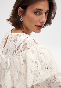 Top en dentelle ivoire avec des motifs floraux texturés, des manches froncées et un dos à ouverture en forme de clé, associé à une boucle d'oreille en argent audacieuse.