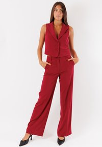 Gilet rosso su misura con due bottoni, abbinato a pantaloni a gamba larga nello stesso tessuto. Il look è completato da scarpe con tacco nero.