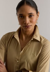 Γυναίκα με σκούρα μαλλιά μαζεμένα πίσω, που φοράει μπεζ πουκάμισο με κουμπιά και μανίκια γυρισμένα και χρυσά σκουλαρίκια σε μονόχρωμο φόντο.