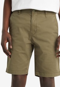 Shorts en coton khaki avec une texture lisse, présentant une fermeture à bouton, des poches latérales et une coupe droite juste au-dessus du genou.