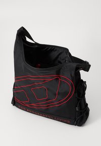 Sac à épaule en nylon noir avec un design brodé rouge, fermeture zippée et sangle réglable. Comprend un cordon de serrage sur le côté et un détail de logo.