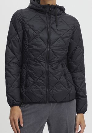 Černá prošívaná bunda s kapucí, předním zipem a zipovanými bočními kapsami, nošená se šedými kalhotami, zobrazena od krku po boky.