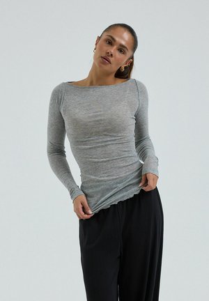 Žena s tmavými vlasmi, oblečená v priliehavom sivom tričku s dlhým rukávom a vo voľných čiernych nohaviciach, jemne si sťahujúca lem trička dole.