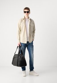 Camicia beige abbottonata sopra una t-shirt bianca, jeans blu, sneakers bianche e tiene una borsa a tracolla nera con logo visibile. Occhiali da sole neri.