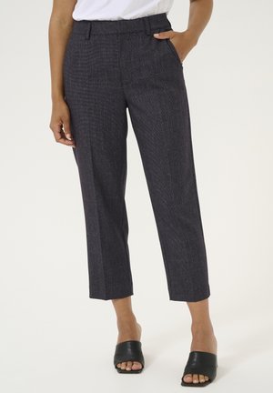 Pantalones oscuros de cuadros con un corte ajustado, longitud recortada y bolsillos laterales. Hechos de tejido texturizado. Combinados con sandalias negras de tipo slide.