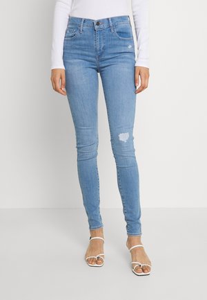 Jean slim en denim bleu clair, avec une taille haute, des poches arrière et un genou déchiré. Porté avec des sandales blanches à lanières.