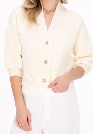 Mujer con un cárdigan color crema abotonado con tres botones dorados y pantalones blancos a juego, posando con una mano cerca de la cintura.