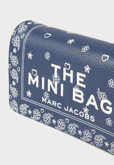 Marineblauw leren mini-tasje met witte bloemen- en hartpatronen, voorzien van gedurfde letters: "THE MINI BAG MARC JACOBS." Gedetailleerd gestructureerd oppervlak.