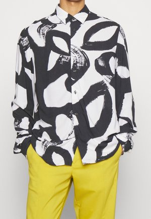 Mężczyzna ubrany w koszulę na guziki z długim rękawem, ozdobioną dużymi abstrakcyjnymi czarno-białymi wzorami, w połączeniu z jaskrawymi żółtymi spodniami. Ręce w kieszeniach.